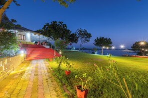 A well-lit outdoor area with a pathway leading to a building with a thatched roof, surrounded by greenery and a view of the water. - Kutch Safari Resort (Bhuj)