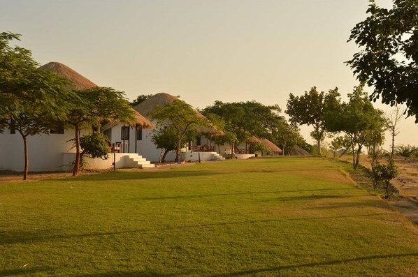 A rural setting with thatched-roof huts and a grassy field. - Kutch Safari Resort (Bhuj)