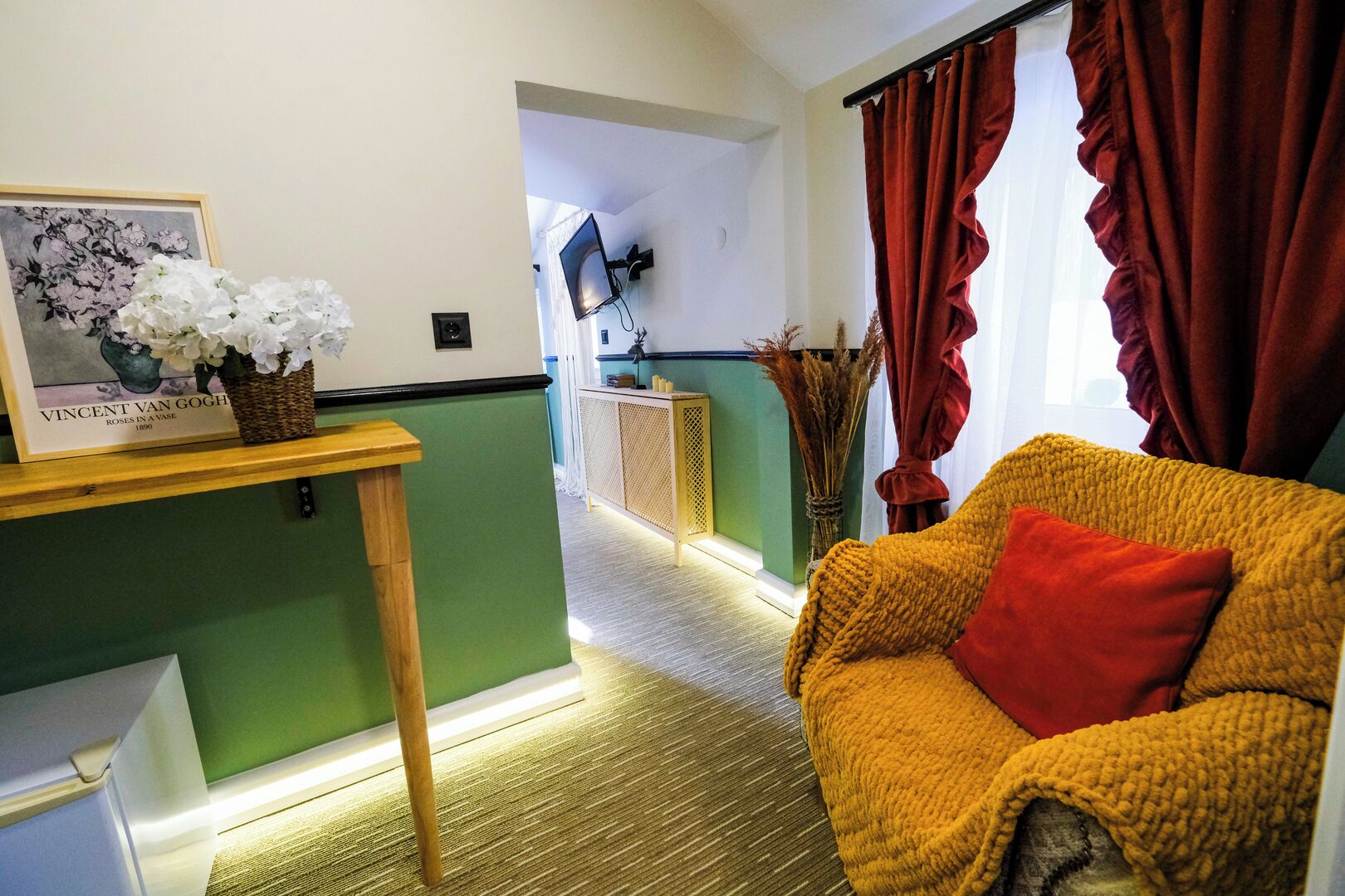 A room with a yellow armchair, a wooden side table with a vase of flowers, a framed picture, and a view of a hallway.