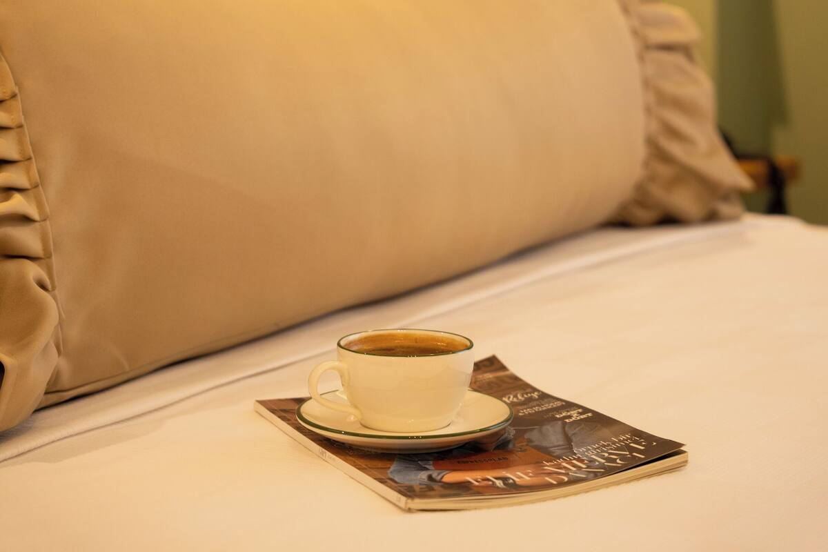 Ein Bett mit einer Tasse Kaffee auf Untersetzer, einer Zeitschrift und einer Wandleuchte.
