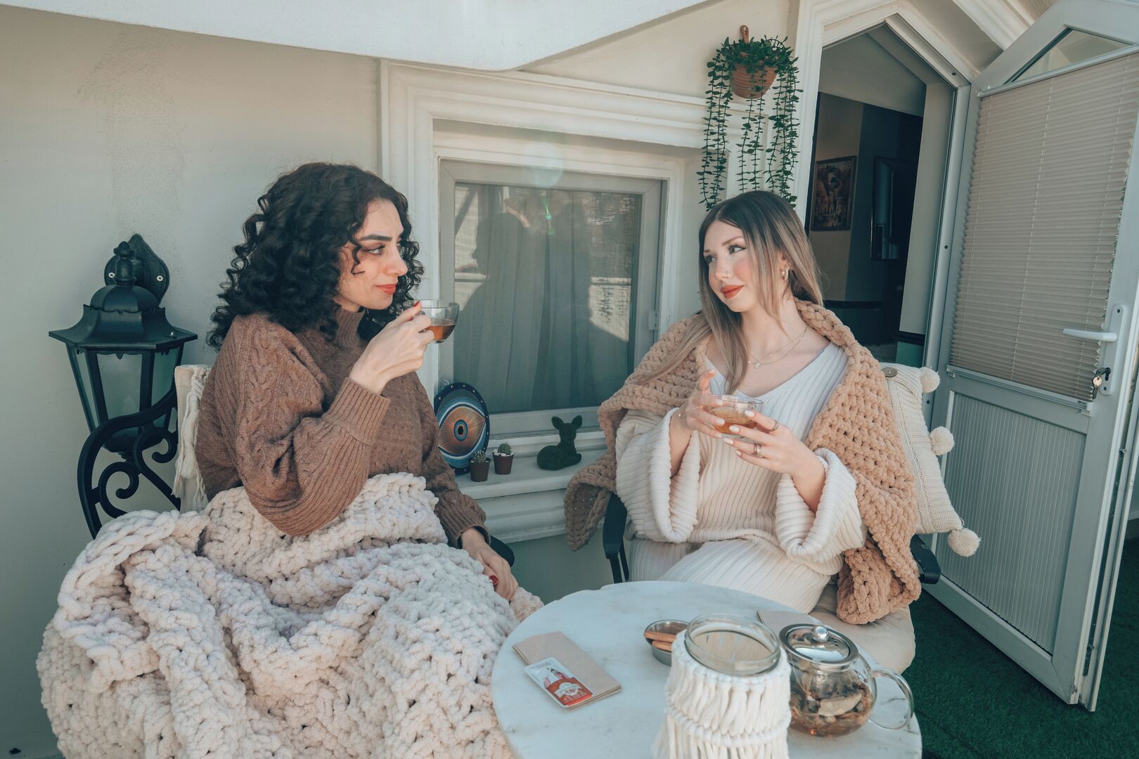 Dos personas sentadas en sillas afuera de un edificio, sosteniendo bebidas, con una mesa frente a ellas.