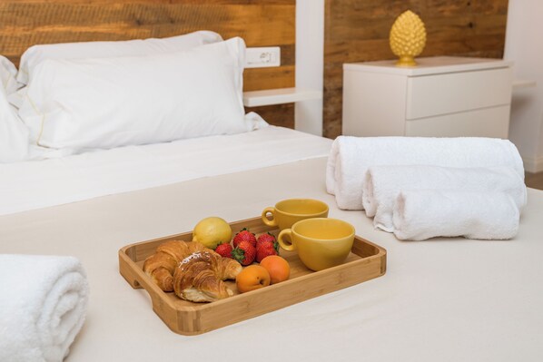 Una cama bien hecha con ropa blanca, una bandeja de madera con cruasanes, frutas y tazas amarillas, y una mesita de noche con una decoración de piña.