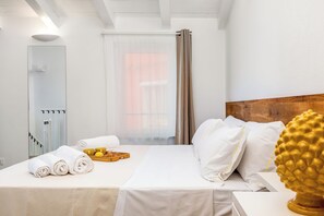 Una habitación de hotel con una cama, cabecera de madera, ventana con cortinas, espejo y una bandeja con toallas y artículos de aseo.