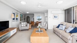 Una sala de estar moderna con un televisor de pantalla plana, una mesa de centro de madera, un sofá color beige y un comedor al fondo.