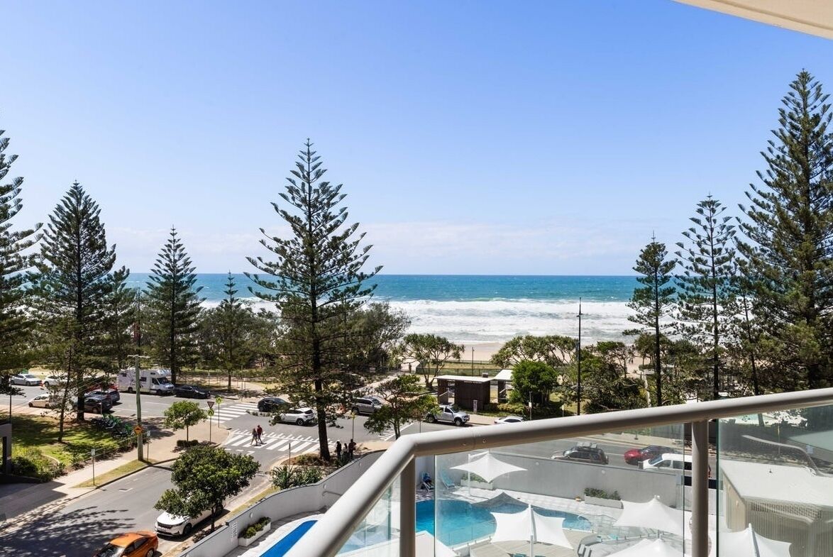 A coastal view from a balcony overlooking a beach, with trees and a pool area visible.