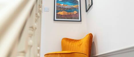 A room with a yellow chair, a framed artwork depicting mountains and a sun, and a white wall.