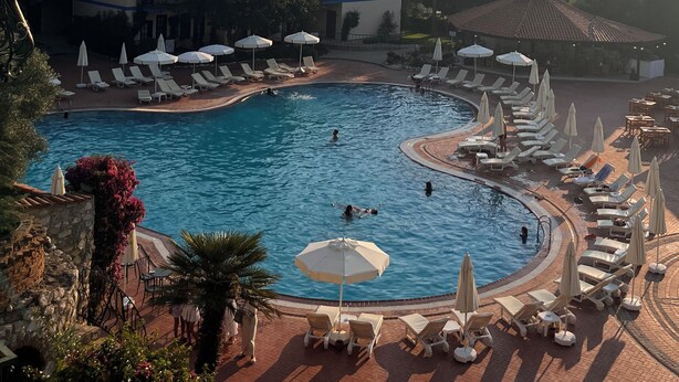Piscina de un complejo turístico con tumbonas y sombrillas, rodeada de edificios y colinas.
