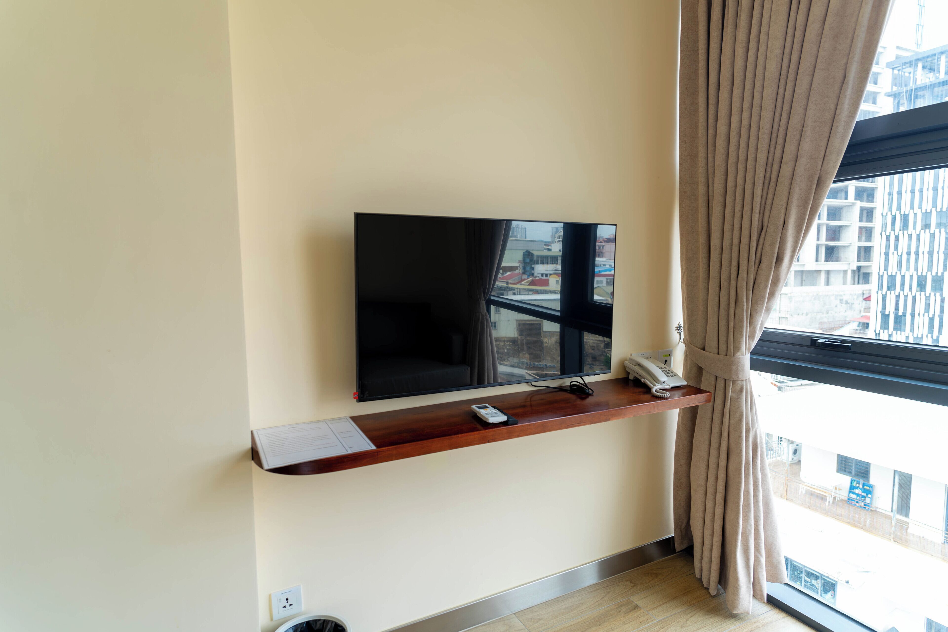 Habitación de hotel moderna con una televisión de pantalla plana montada en un estante de madera, un teléfono y una ventana con cortinas.