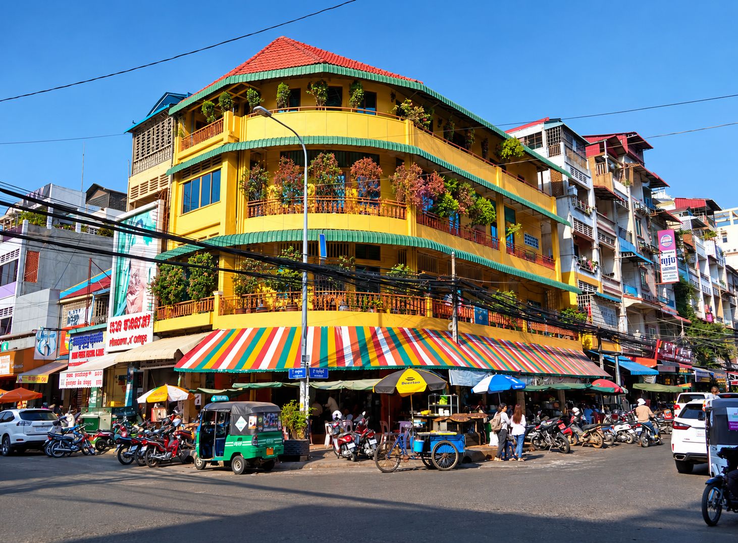 Un edificio de varios pisos con balcones, una tienda en la planta baja con toldos de colores, y una calle con motocicletas estacionadas y un tuk-tuk.