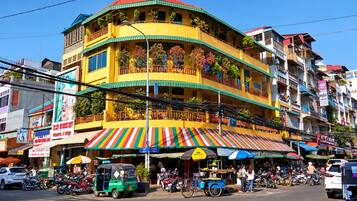 Un edificio de varios pisos con balcones, una tienda en la planta baja con toldos de colores, y una calle con motocicletas estacionadas y un tuk-tuk.