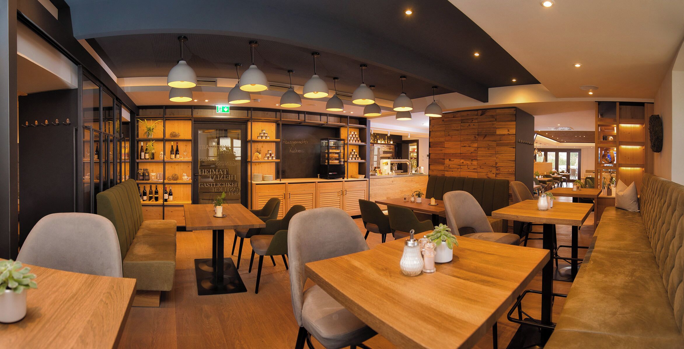 Un interior de restaurante moderno con mesas de madera, asientos acolchados, iluminación colgante y una zona de bar con estantes llenos de botellas.