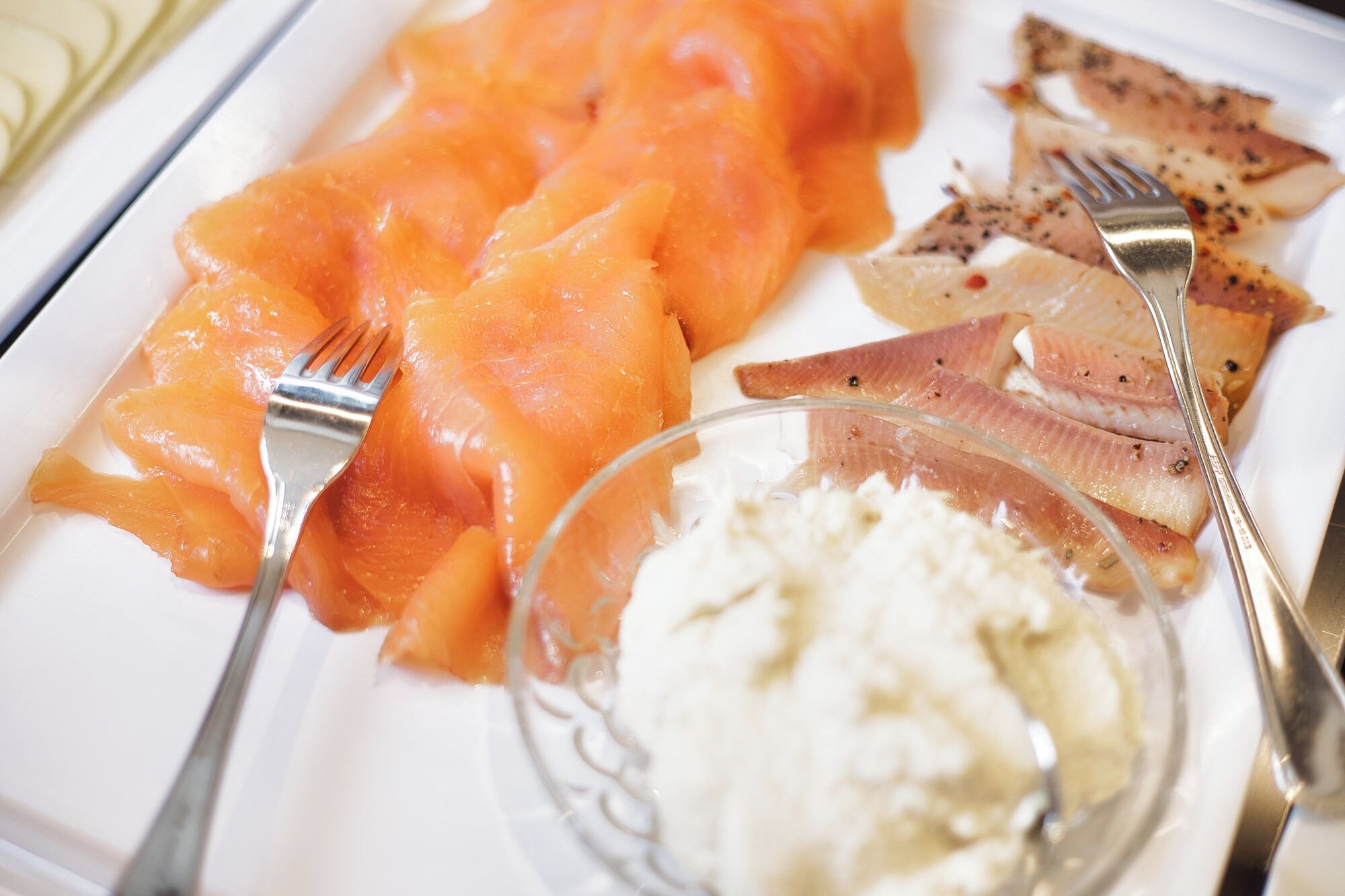 A plate with smoked salmon, pickled herring, and a small bowl of cream cheese.