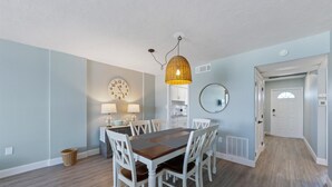 Un comedor con una mesa de madera y sillas blancas, una lámpara colgante, un reloj de pared y un espejo.