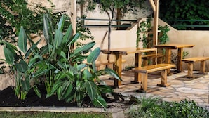 An outdoor patio with wooden tables and benches, surrounded by lush green plants and a stone floor. - Konstantinos Hotel & Apartments I (Nidri)