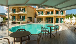 Uma área de piscina com espreguiçadeiras e mesas, um espaço coberto para sentar e um edifício com varandas.