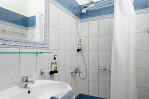 Casa de banho com lavatório branco, chuveiro com prateleira de vidro e espelho com azulejos decorativos.
