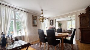 A dining area with a wooden table, black chairs, and a patterned rug. There is a chandelier, a clock on the wall, and a window with curtains. - Charming and spacious villa near Lidköping |SE08085 (Lidköping)