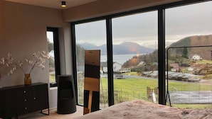 Un dormitorio con una cama grande, un aparador, un jarrón con flores y vistas a un lago y montañas.