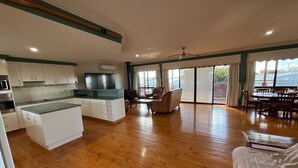 A spacious living area with a kitchen, wooden flooring, a dining table, and a large window with curtains.