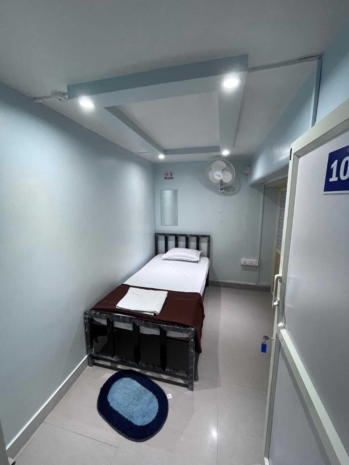 A bedroom with a bed, a bedside table, a wall-mounted fan, and a door with a number sign.