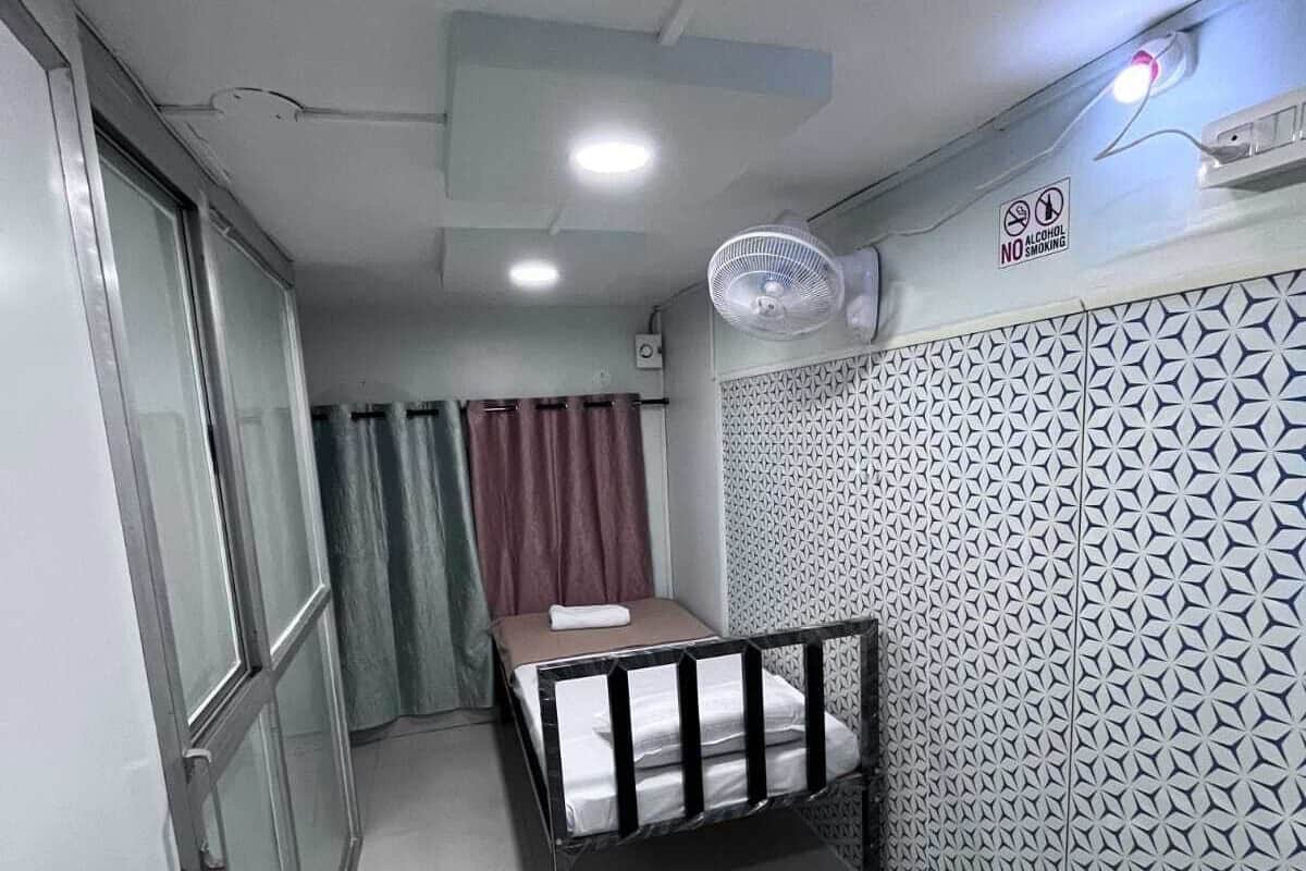 A small, compact room with a bed, a window, a door, and a wall-mounted air conditioner.