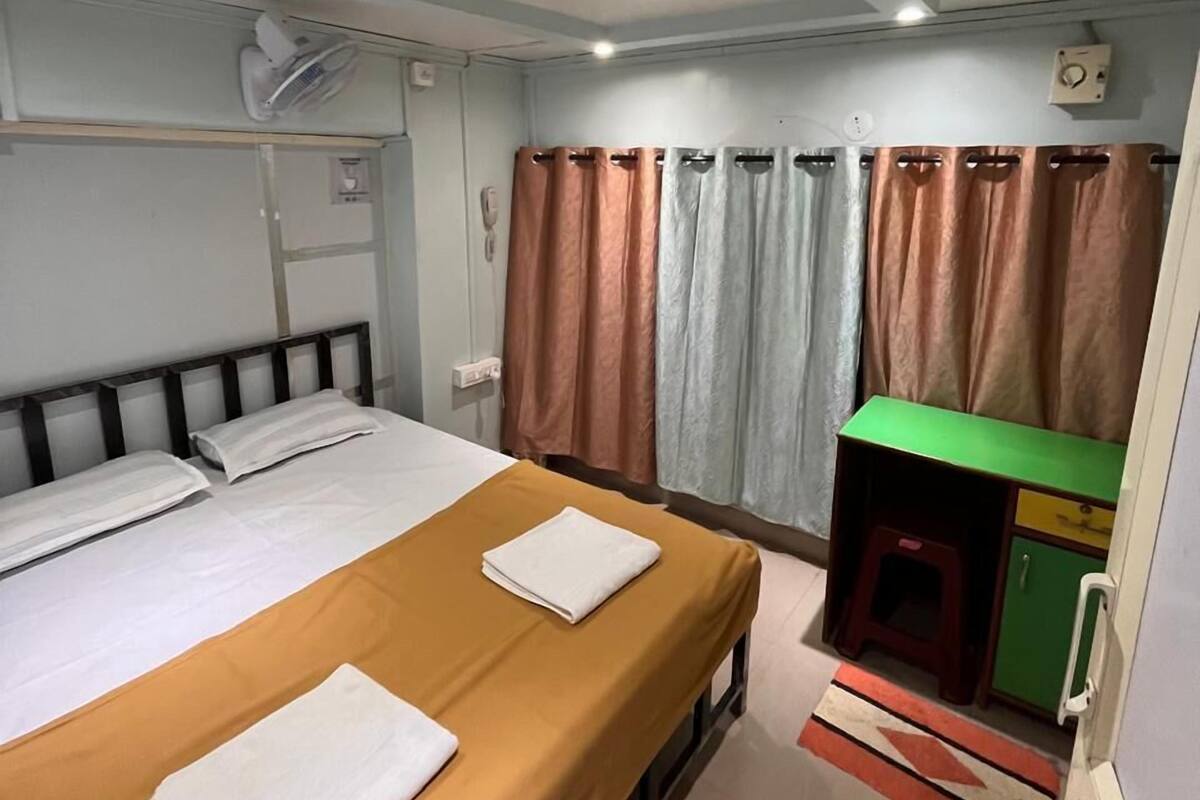 A hotel room with two beds, a green desk, a small cabinet, and a wall-mounted air conditioner.