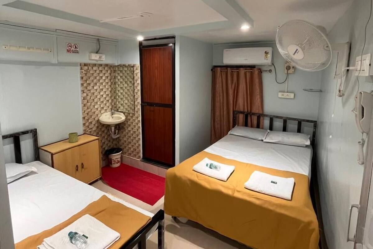 A compact hotel room with two beds, a small desk, a bedside table, a fan, and an air conditioning unit.