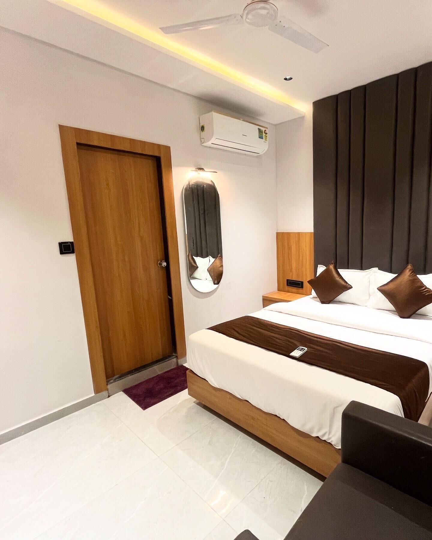 A modern bedroom with a wooden door, a bed with white and brown bedding, a bedside table with a lamp, and a ceiling fan.