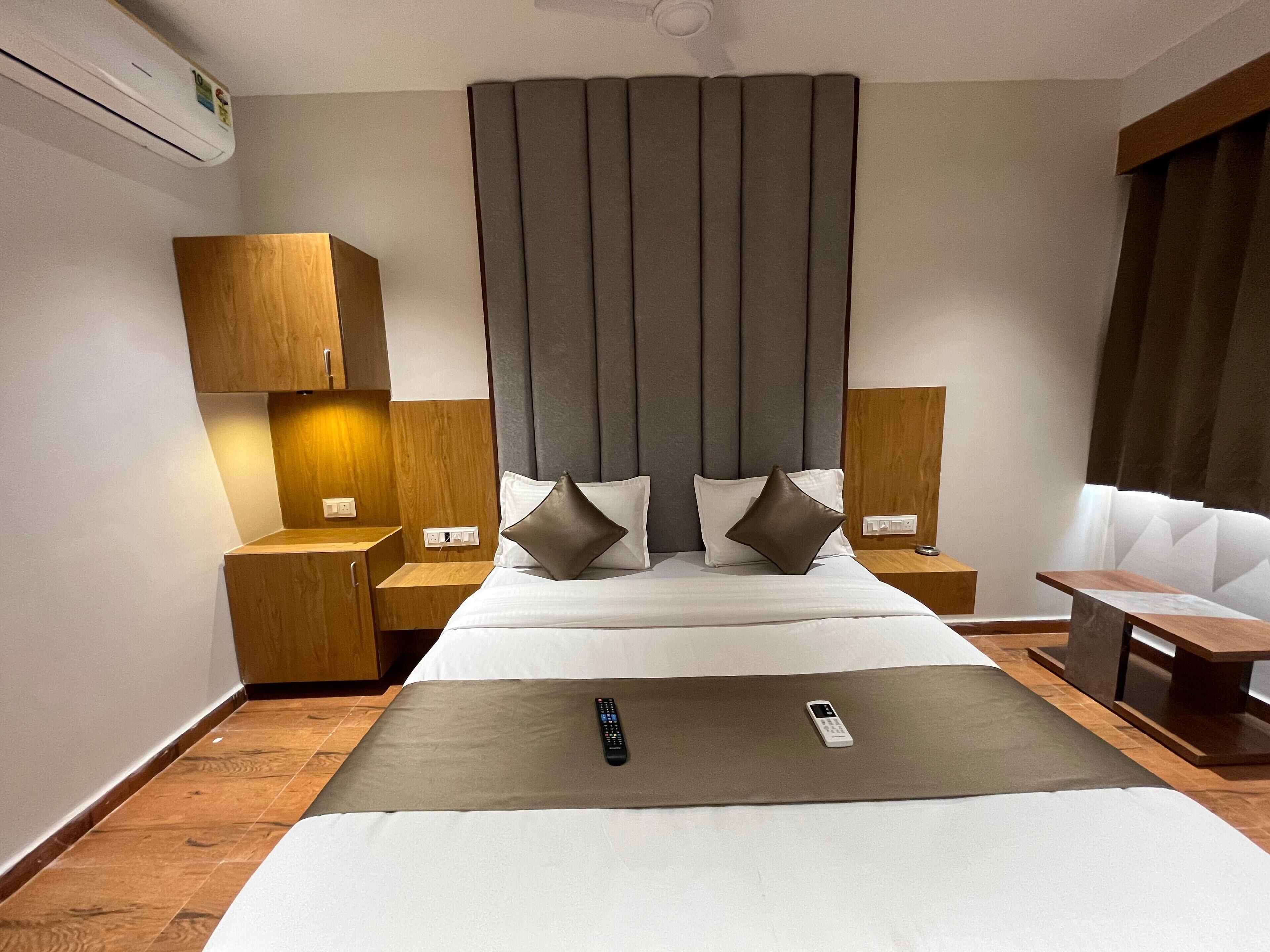 A modern hotel room with a large bed, a bedside table with a lamp, a small bedside table with a lamp, a remote control on the bed, and a chair with a table.
