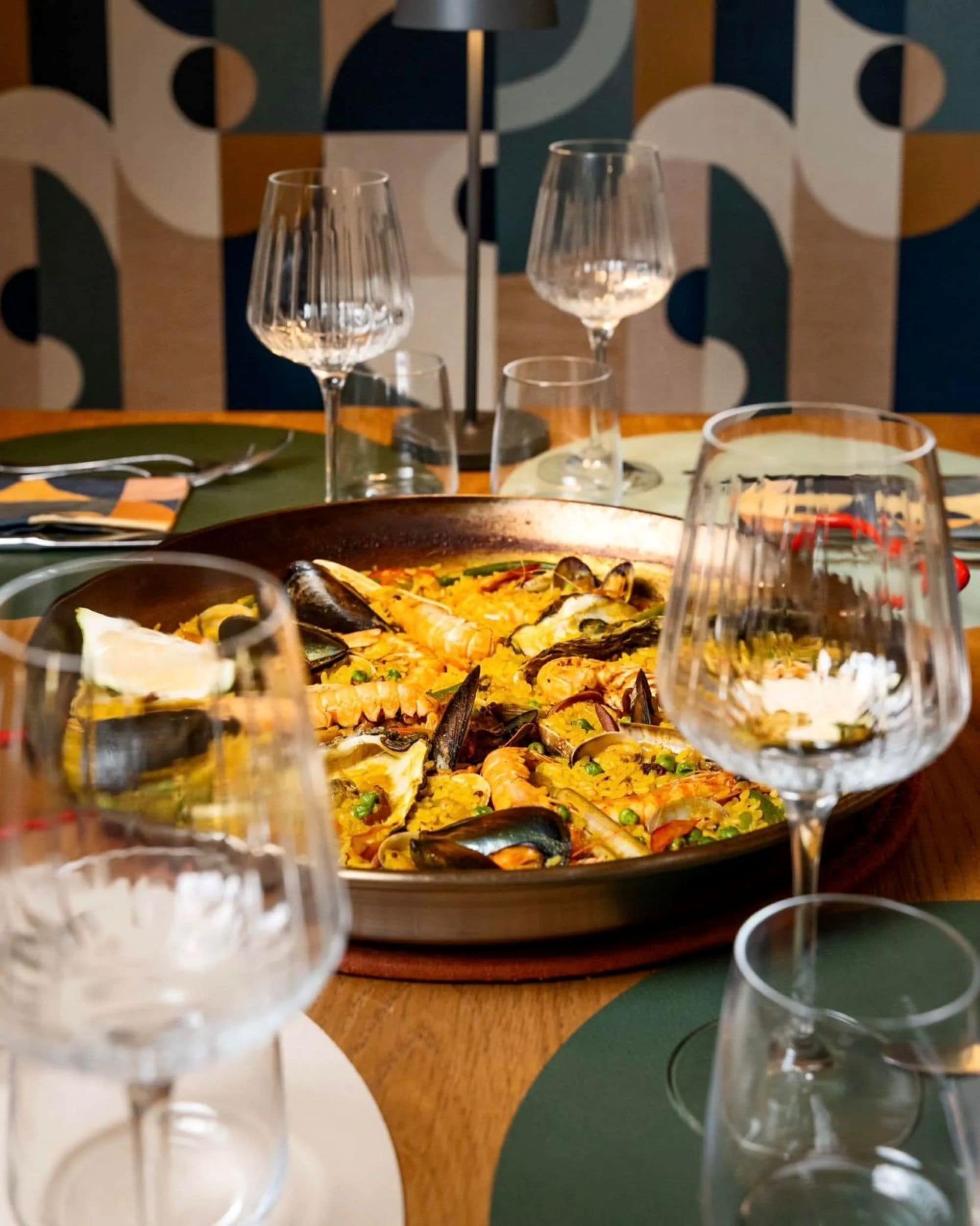 Una mesa de comedor con una paella de mariscos, copas de vino y un papel tapiz estampado al fondo.