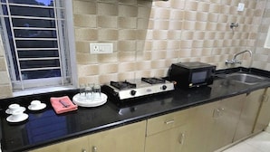 A kitchen with a gas stove, a microwave, and a sink. There is a window with blinds and a tiled backsplash. - Brindavan Service Apartments (Mysore)