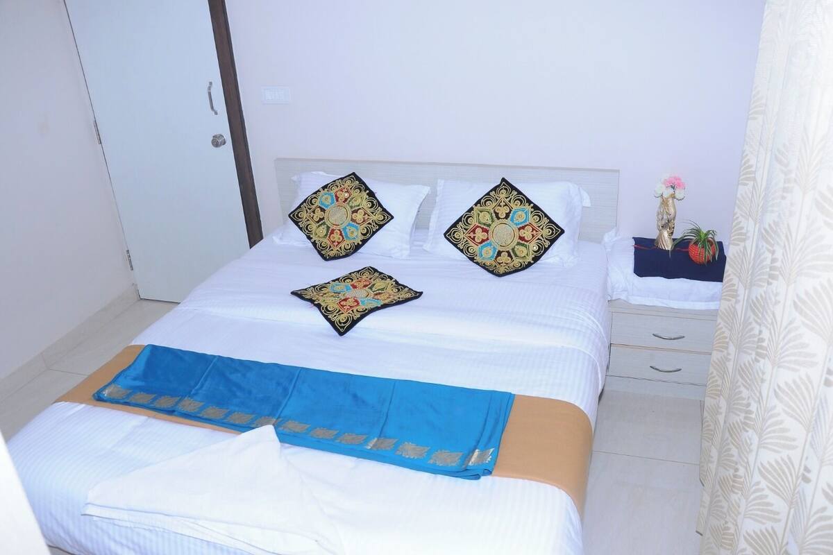 A neatly made bed with white linens and decorative pillows, a blue bedspread, and a small bedside table with a plant.