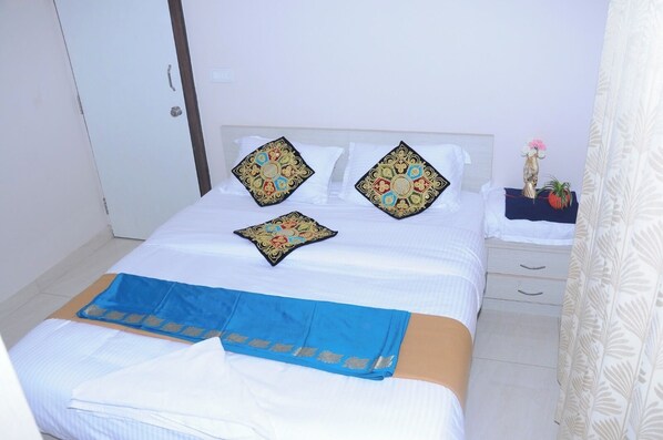 A neatly made bed with white linens and decorative pillows, a blue bedspread, and a small bedside table with a plant. - Brindavan Service Apartments (Mysore)