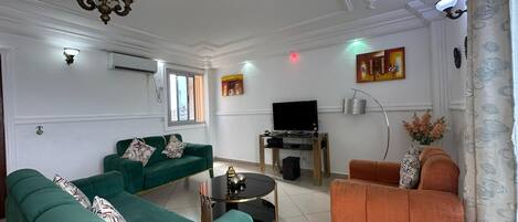 Una sala de estar con dos sofás verdes, un sillón naranja, un televisor sobre un mueble bajo y una araña.