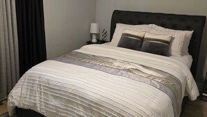 Een netjes opgemaakt bed met een wit en geborduurd dekbed, een donker hoofdbord en een nachtkastje met een lamp en een kleine plant.