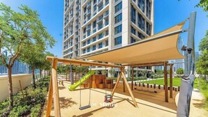 Un terrain de jeux avec des balançoires et un toboggan, entouré d’un parc arboré, avec un grand immeuble en arrière-plan.