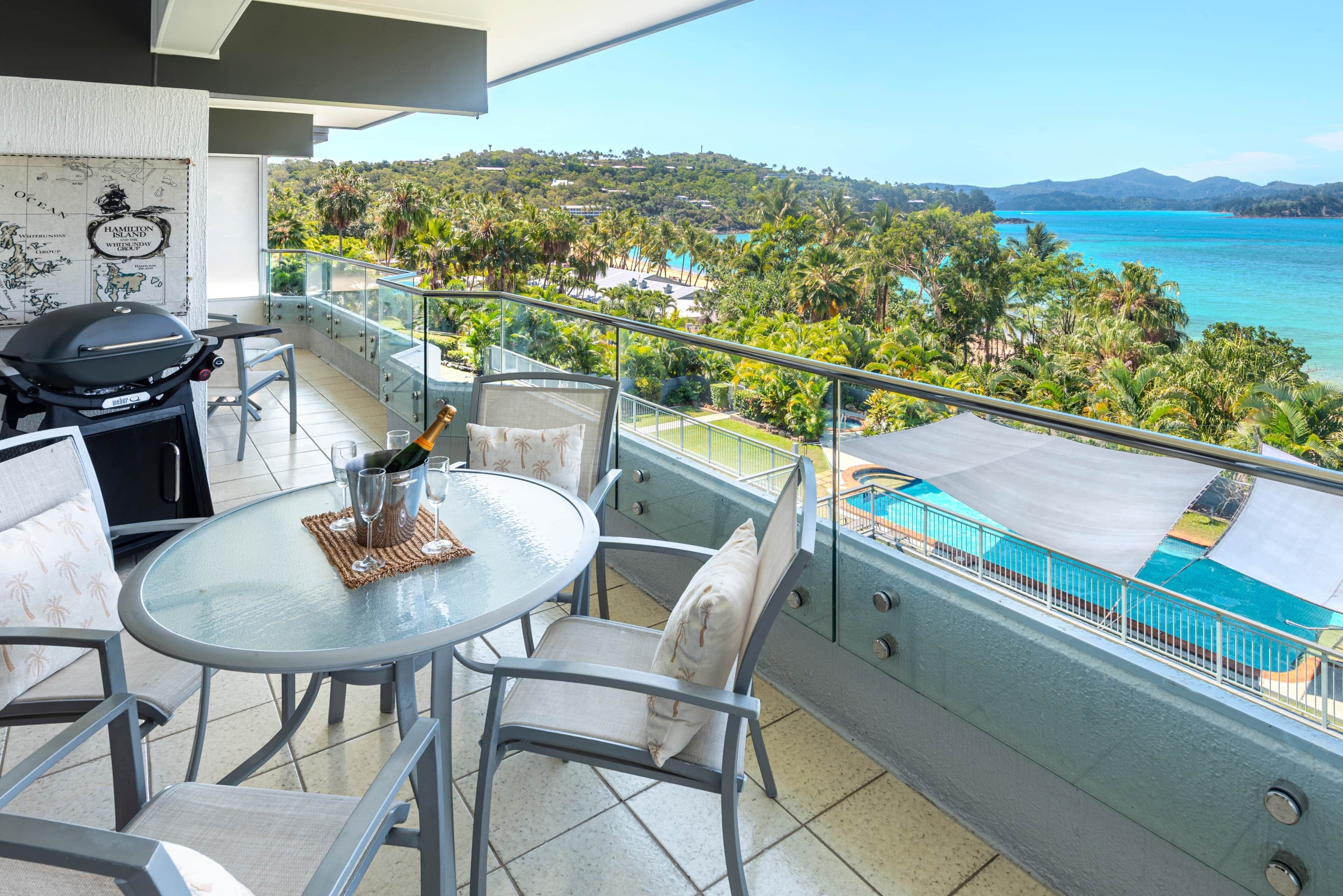A balcony with a glass table, chairs, and a grill. It overlooks a pool and a tropical landscape.