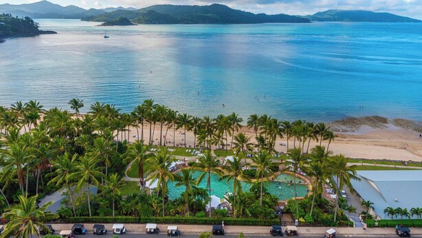 Un resort en la playa con piscina, palmeras y vistas al océano y las montañas.