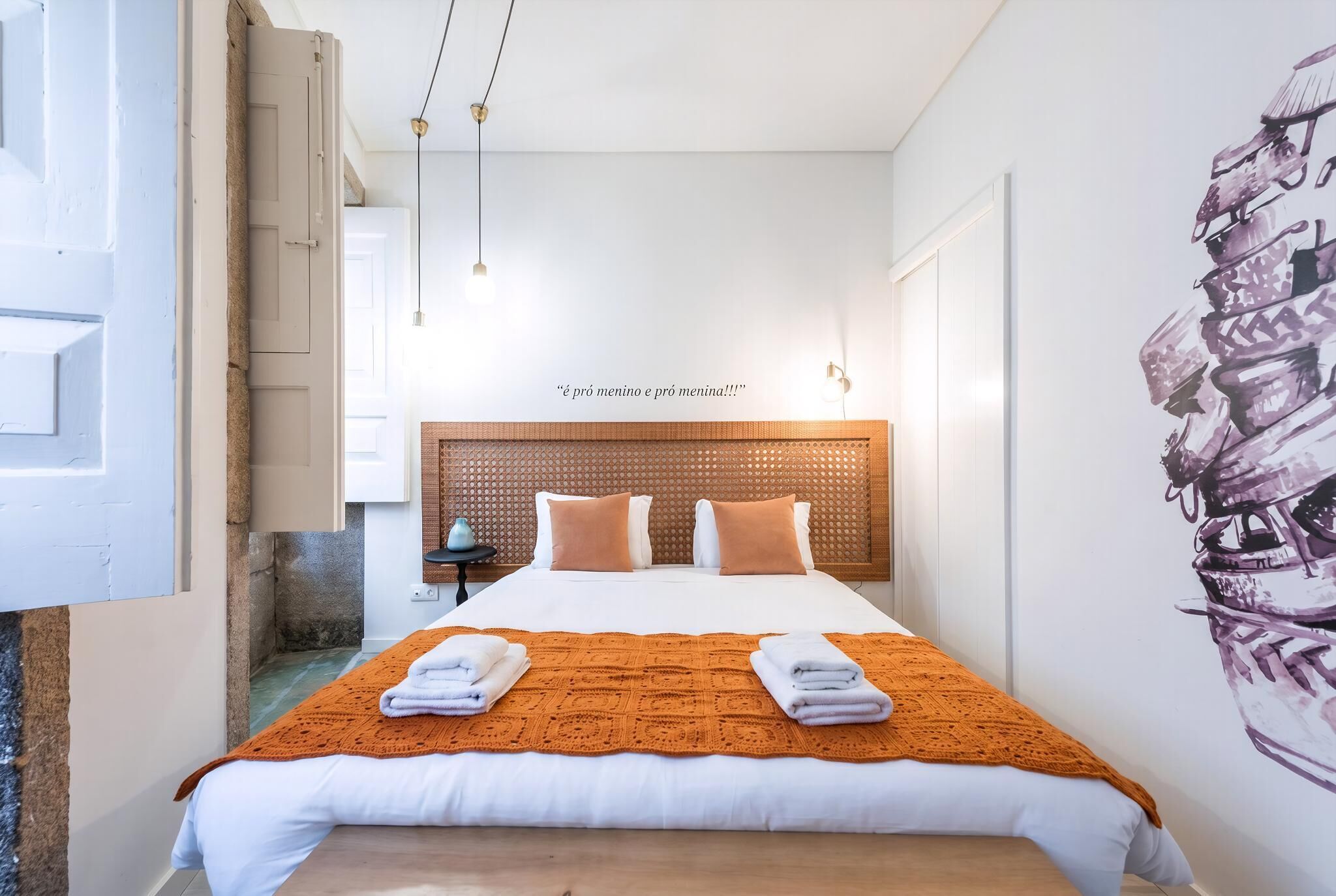 Un lit bien fait, recouvert d’une couverture à motifs, avec une tête de lit ornée d’un motif tissé, et un mur décoré d’un motif graphique représentant des valises empilées.