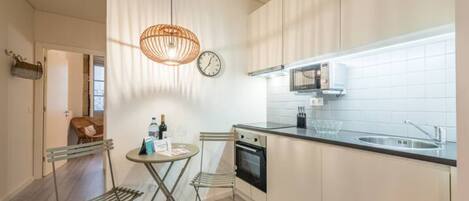 A modern kitchen with a dining area, featuring a round table, two chairs, a hanging light fixture, a clock, and a built-in oven.