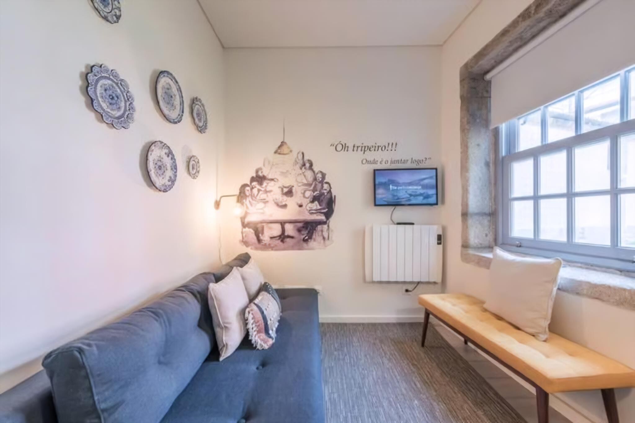 Una habitación con un sofá azul, un banco de madera, placas decorativas, un televisor de pared y una ventana.