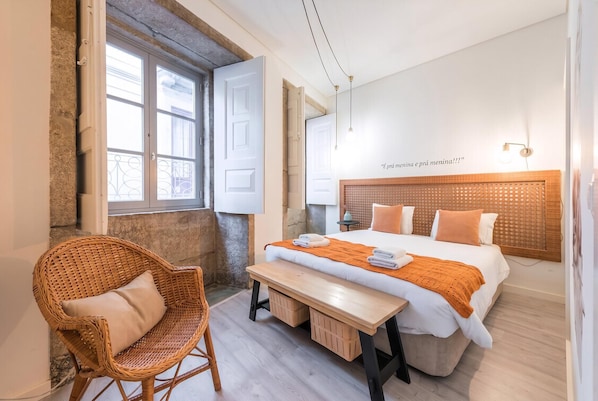 A bedroom with a bed, a bench, a wicker chair, and a window.
