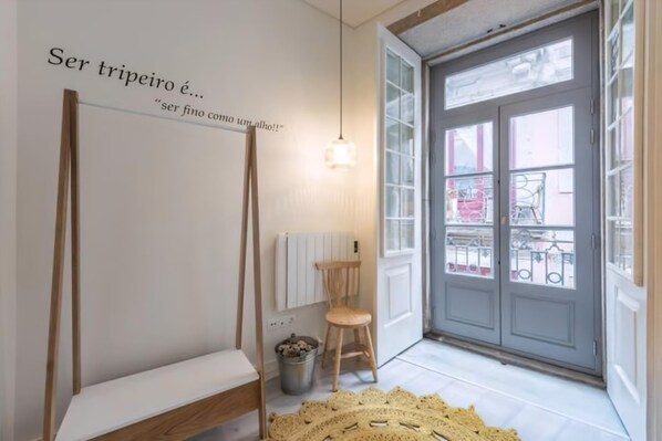 A room with a white wall, a wooden bench, a chair, a radiator, a small trash bin, and a large window with a view of the outside. - Guestready - Genuine Residences (Porto)