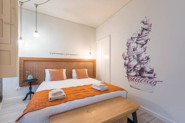 Un dormitorio con cabecera de madera, cama con ropa blanca, manta naranja, un banco y un mural en la pared.