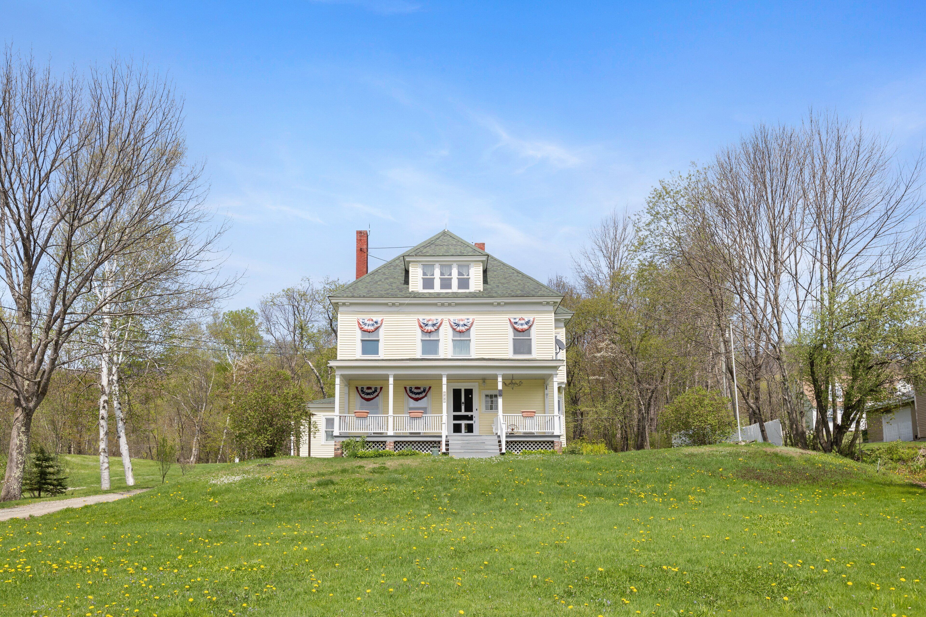 A two-story house with a front porch, a chimney, and a lawn with yellow flowers.