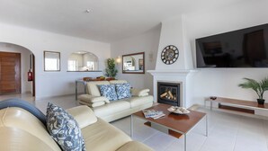 A modern living room with a beige sofa, a coffee table, a fireplace, a TV mounted on the wall, and a dining area in the background. - Apartment do Ramses with oceanviews (Luz)