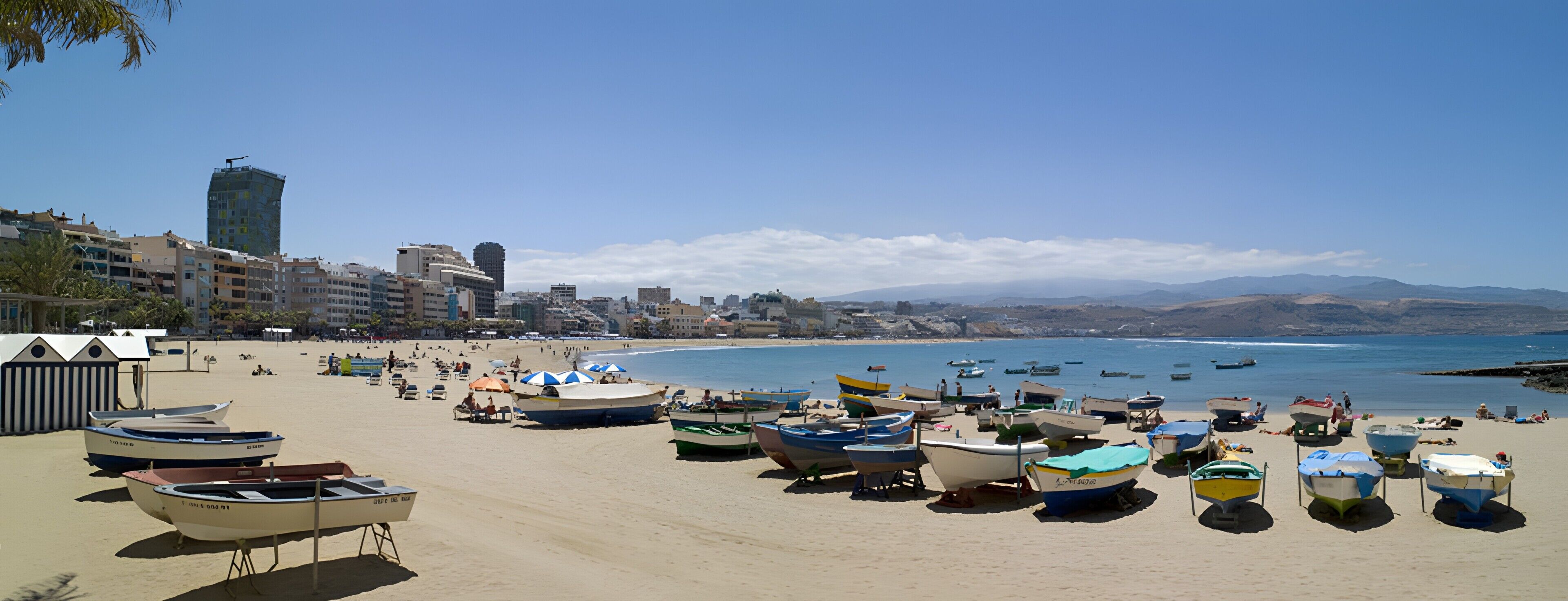 A beach with boats, a clear sky, and buildings in the background.