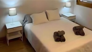 Una cama bien hecha con almohadas, dos mesitas de noche con lámparas y dos toallas enrolladas sobre la cama.