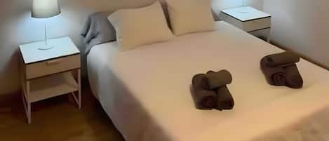 Uma cama bem arrumada com almofadas, duas mesinhas de cabeceira com abajures e duas toalhas enroladas sobre a cama.