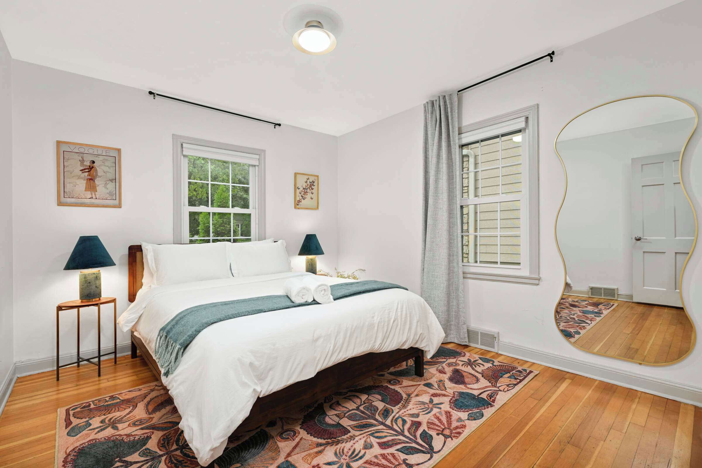 Une chambre à coucher avec un lit, deux lampes de chevet, un grand miroir et une fenêtre avec des stores.